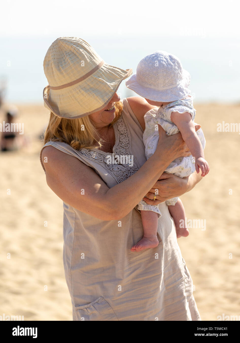 Grandmother holding a baby, both wearing sunhats for protection on a hot and sunny day on a beach. Stock Photo