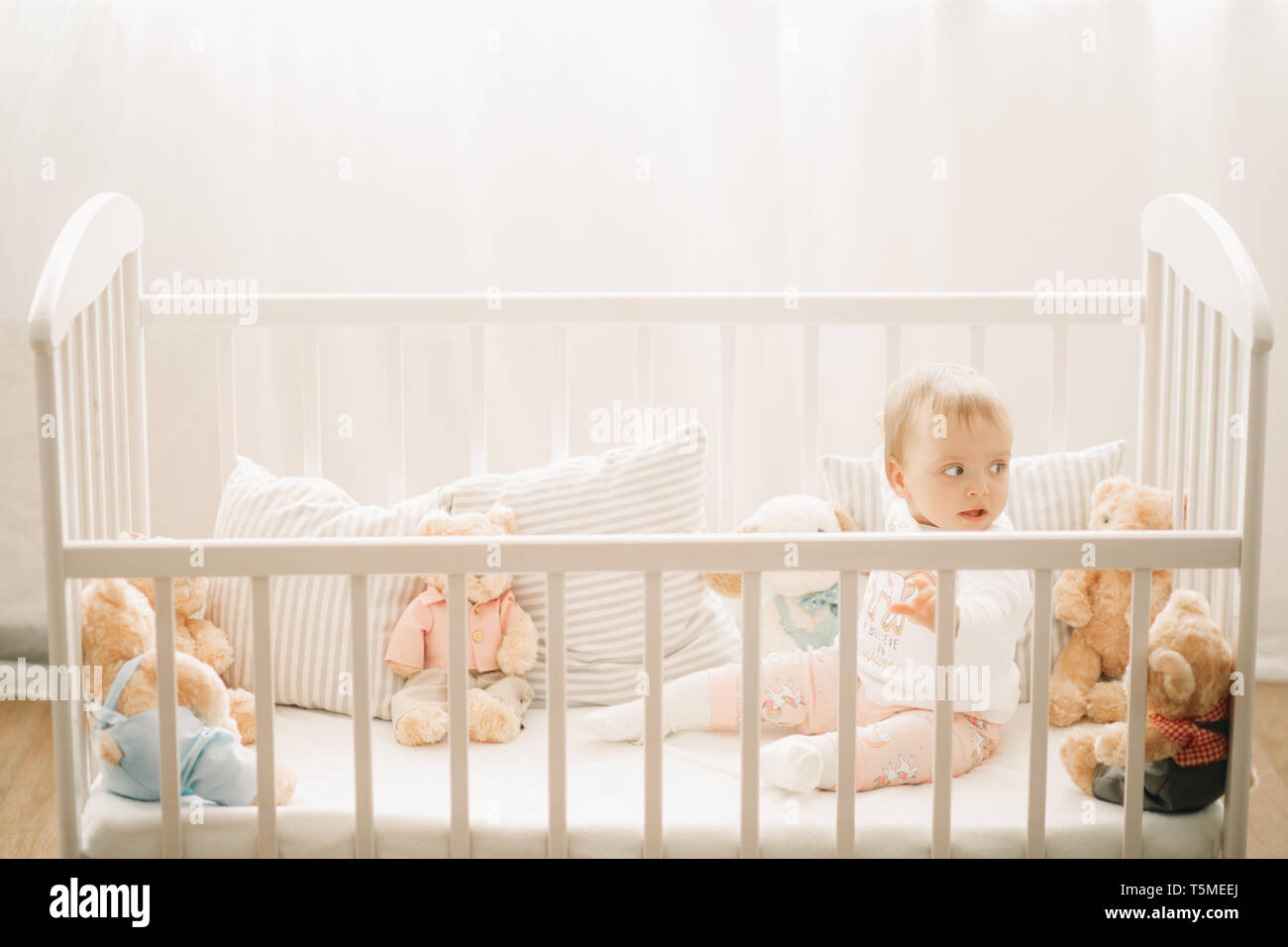 The Toddler Sits In A Crib And Plays Among Pillows And Teddy Bears