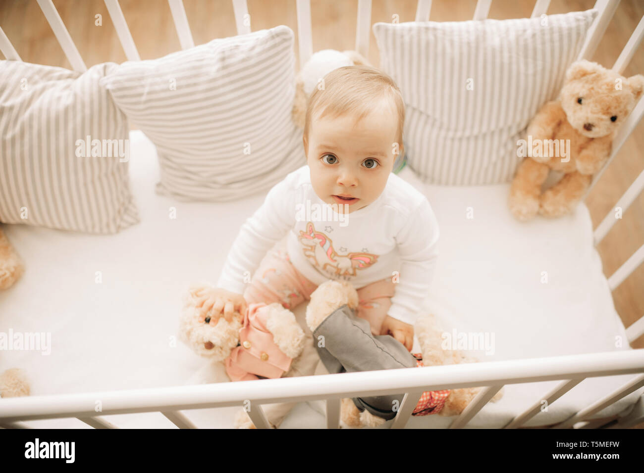 The Toddler Sits In A Crib And Plays Among Pillows And Teddy Bears