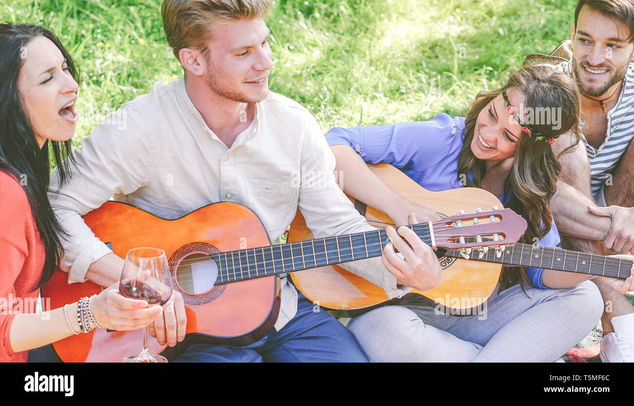 Group of friends playing guitars and singing while drinking red wine sitting on grass in a park outdoor - Happy people having fun together Stock Photo