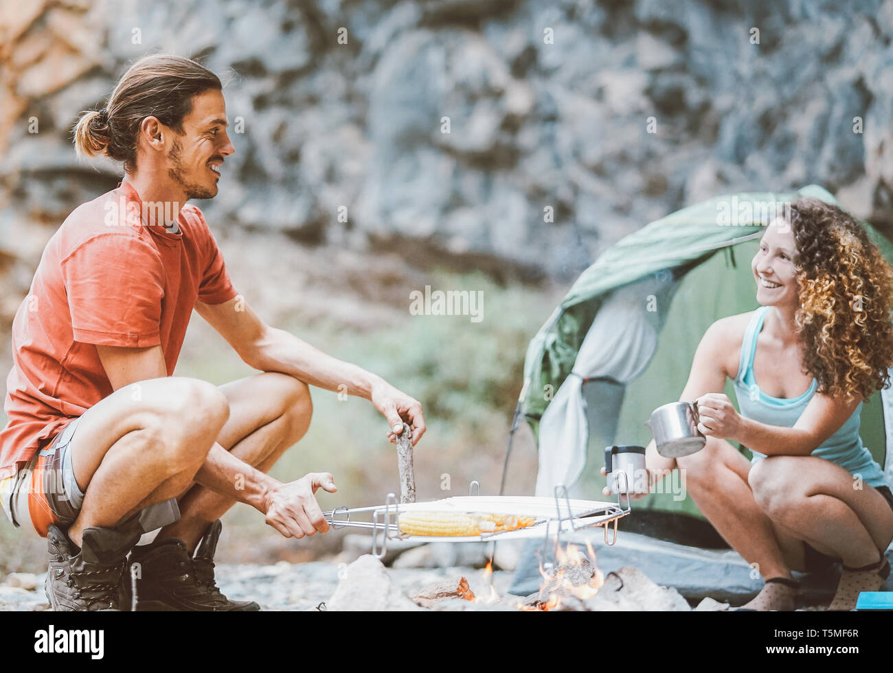 Trekker couple camping in rock mountains with a tent - Climber people cooking and drinking hot tea next to bonfire Stock Photo