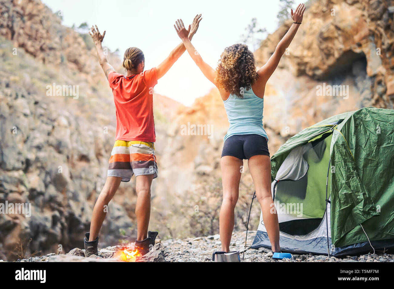 Health young couple doing yoga next to fire while camping with tent on a mountain - Friends meditating together spreading their hands up Stock Photo