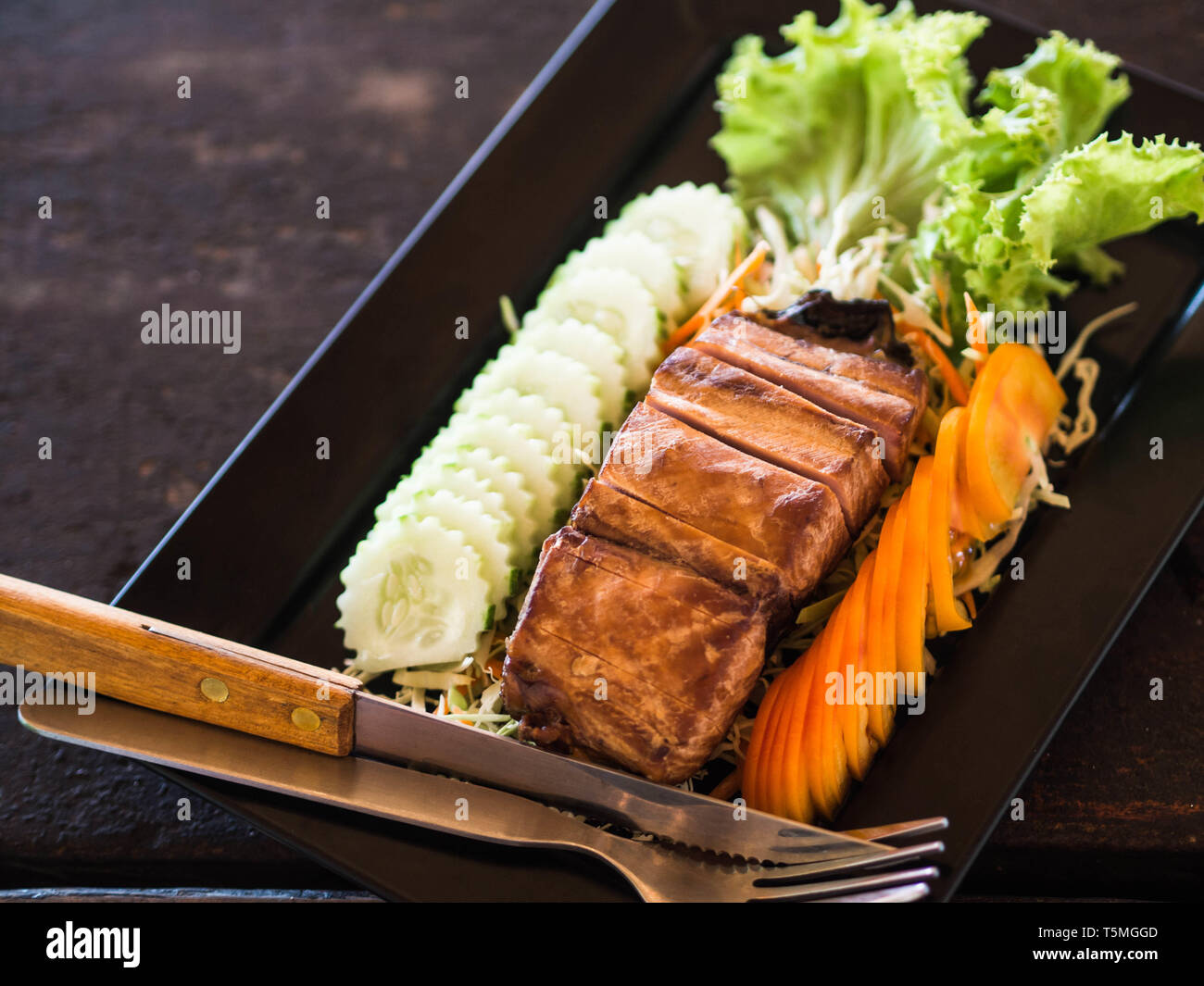 A piece of smoked sea fish with slices of fresh vegetables and cutlery on a black rectangular plate on a wooden table in a Thai restaurant. Stock Photo