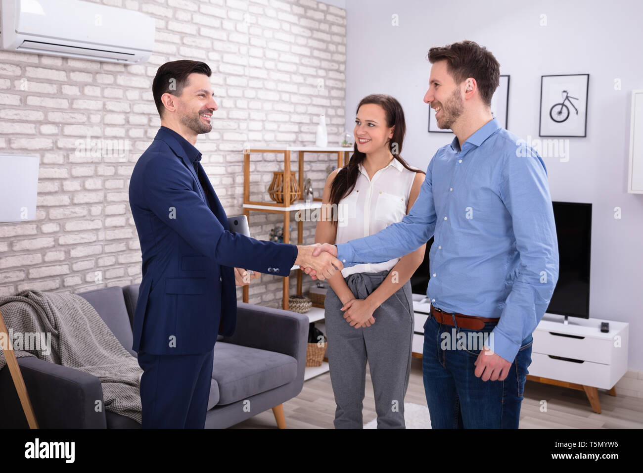 Smiling Male Real Estate Agent Shaking Hands With His Clients In New Home Stock Photo