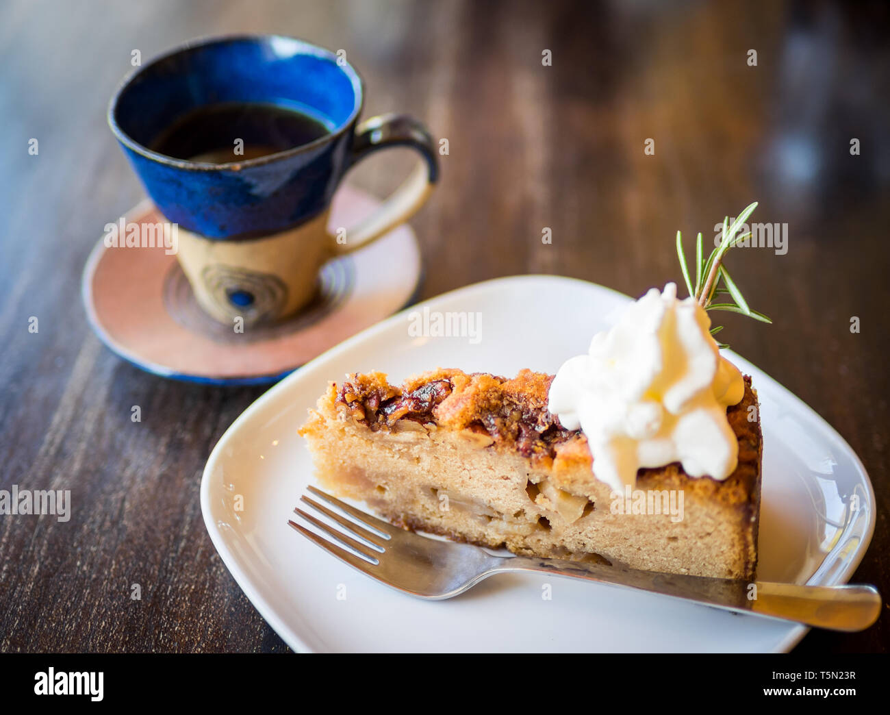 A slice of apple cake with whipped cream and a sprig of rosemary, and a cup of tea, at the tearoom at Westholme Tea Farm near Duncan, BC, Canada. Stock Photo