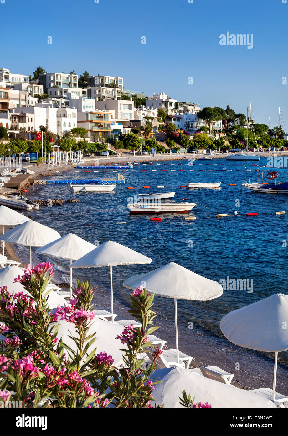 White Umbrellas and sunbeds near lagoon with boats on the beach in Bodrum, Turkey Stock Photo