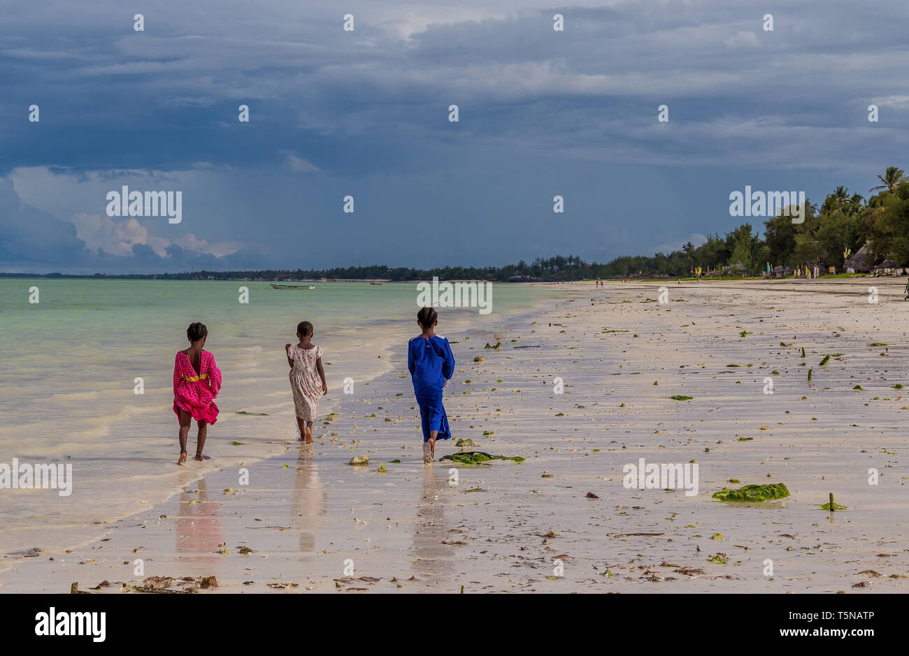 Zanzibar, Tanzania, march 23, 2018 Three little African girls in colorful and bright dresses walk barefoot along the ocean at sunny day. Stock Photo