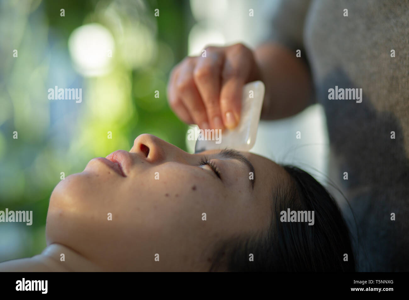 Young woman receiving facial rejuvenation gua sha treatment with rose quartz in acupuncture wellness spa clinic Stock Photo
