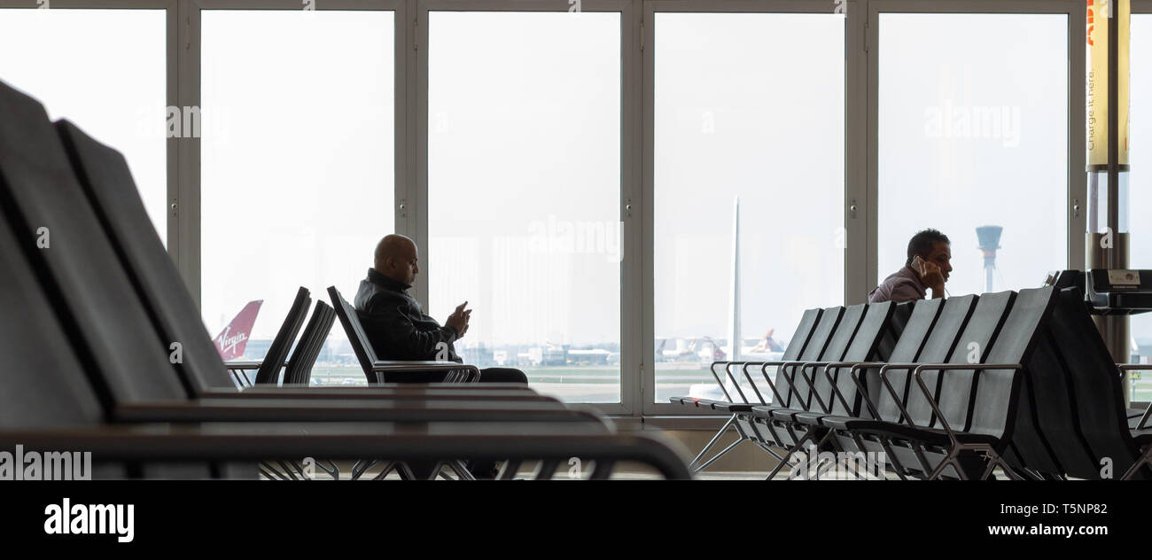 Terminal 4,Heathrow Airport, London, UK April 22 2019. Two men using smart phones are seated at a gate in the airport waiting to board a flight Stock Photo