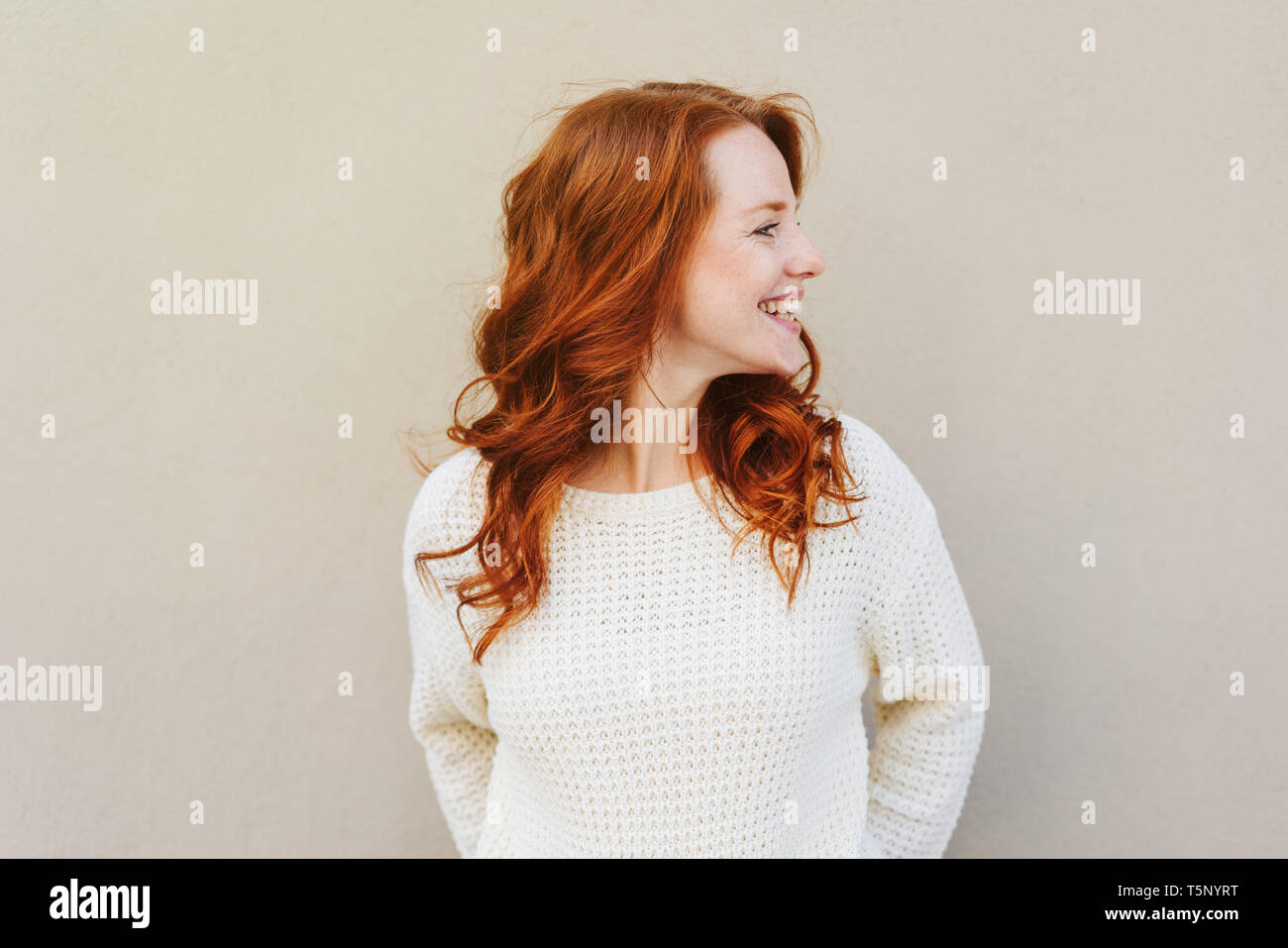 Front portrait of young red-hared woman in white sweater, smiling and looking away, standing against grey wall with her head turn left Stock Photo