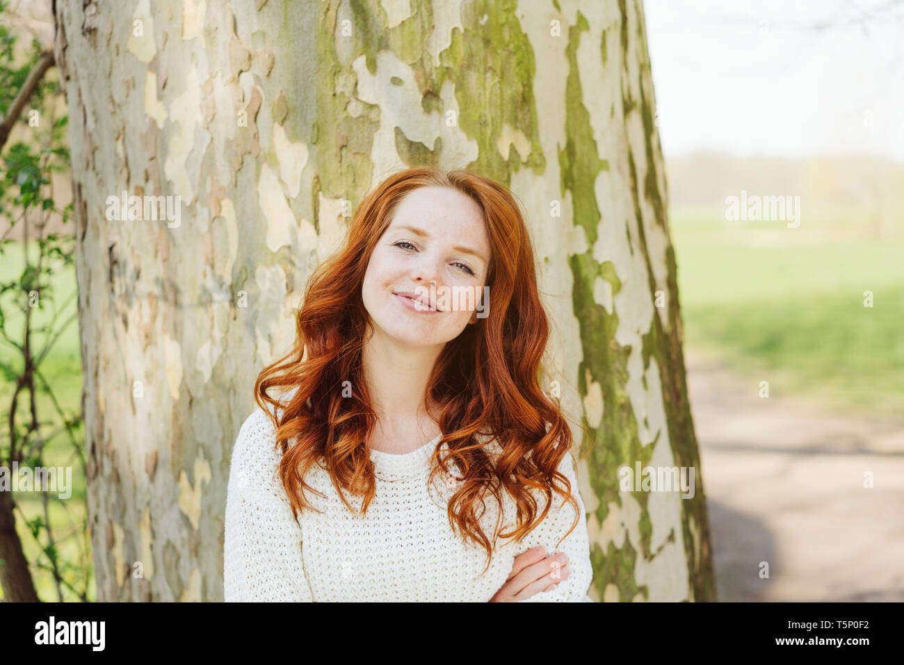 Smiling young woman outdoors in a park standing with folded arms in front of a tree trunk in the warm sunshine Stock Photo