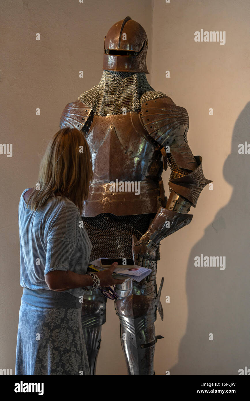A woman checking out an exhibit of a medieval suit of armour, illuminated by lighting from behind her. Belcastel, Occitanie, France. Stock Photo