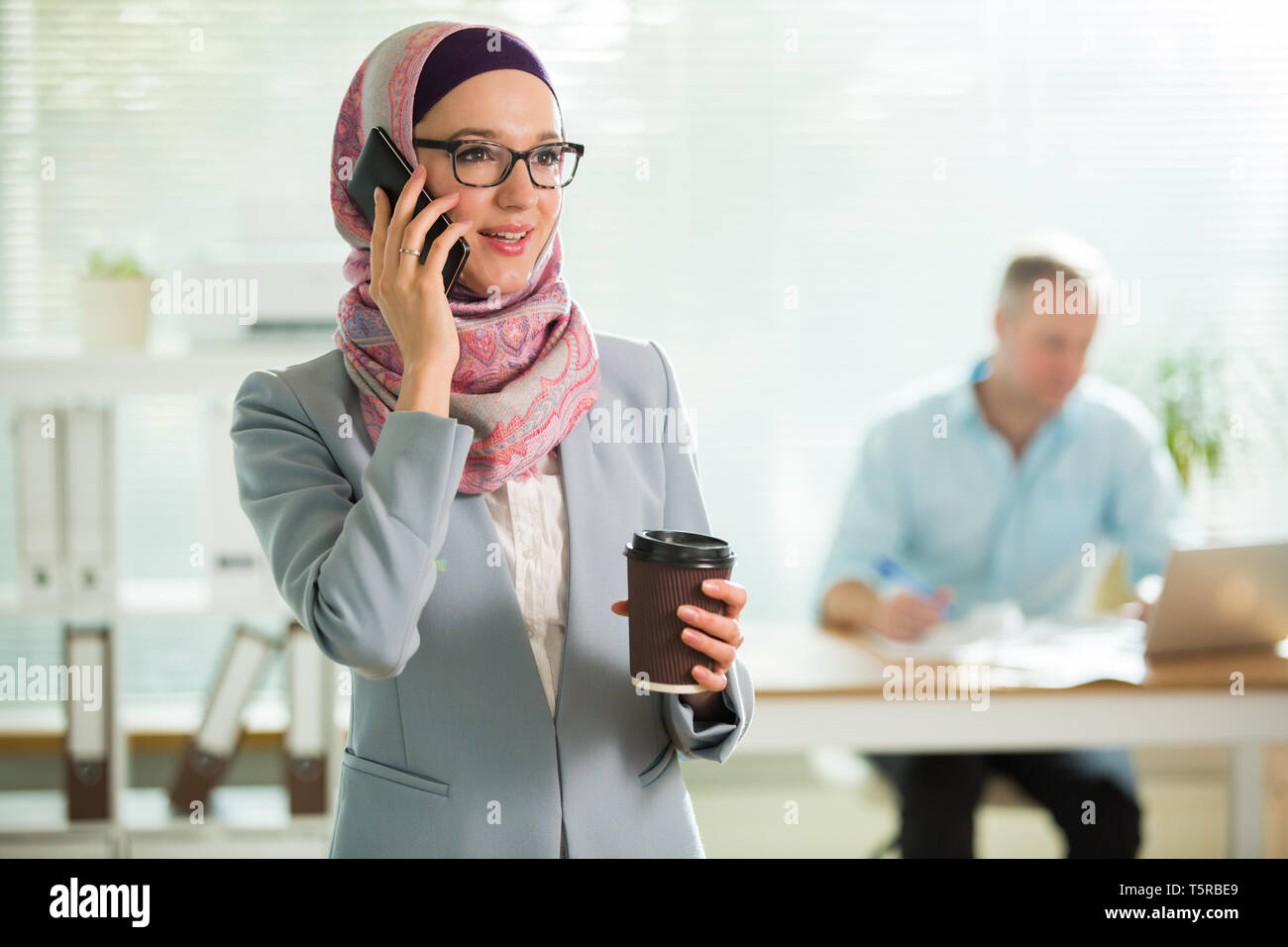 Beautiful woman in hijab and eyeglasses talking on phone, smiling, holding cup of coffee. Portrait of confident muslim businesswoman. Modern office Stock Photo