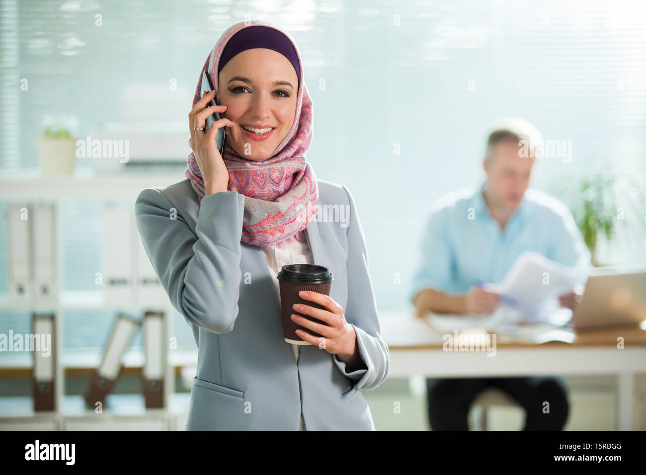 Beautiful woman in hijab and eyeglasses talking on phone, smiling, holding cup of coffee. Portrait of confident muslim businesswoman. Modern office Stock Photo