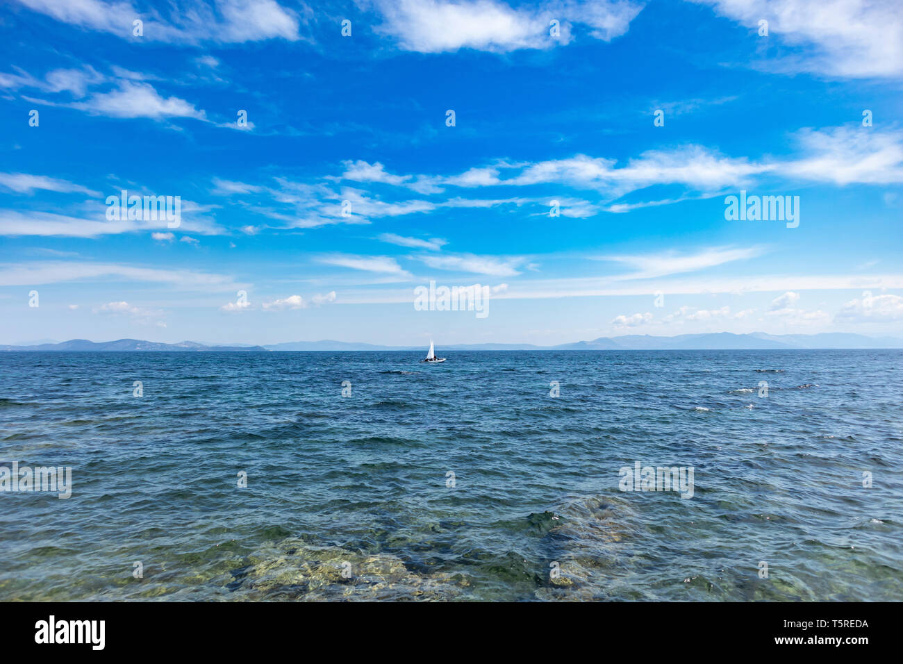 Sailing in Aegean sea, Greece. Small optimist boat with white sail, blue sky and sea background Stock Photo