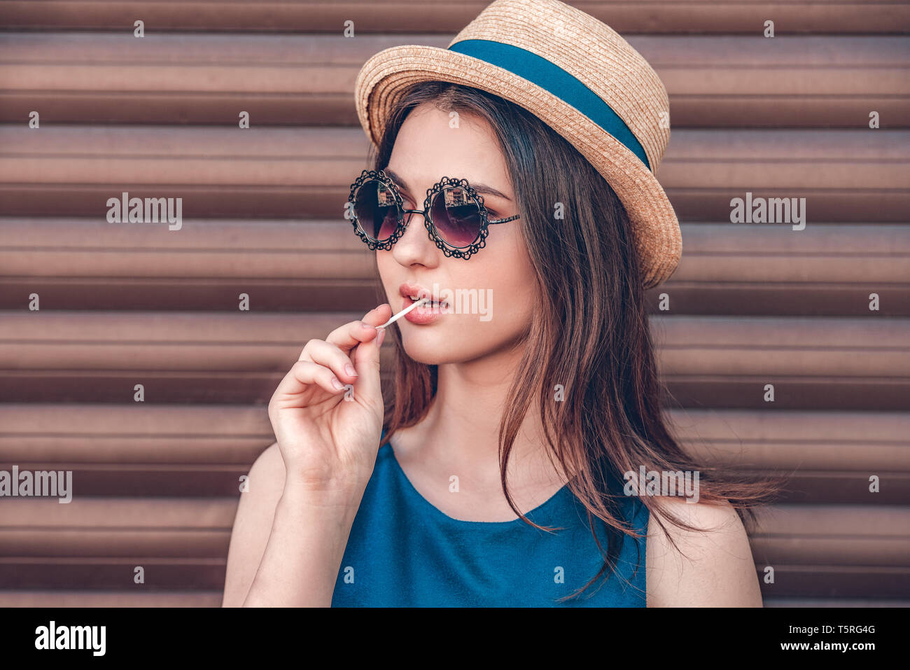 Teenager girl wearing hat and sunglasses standing isolated on wall eating  candy looking aside cool face close-up Stock Photo - Alamy