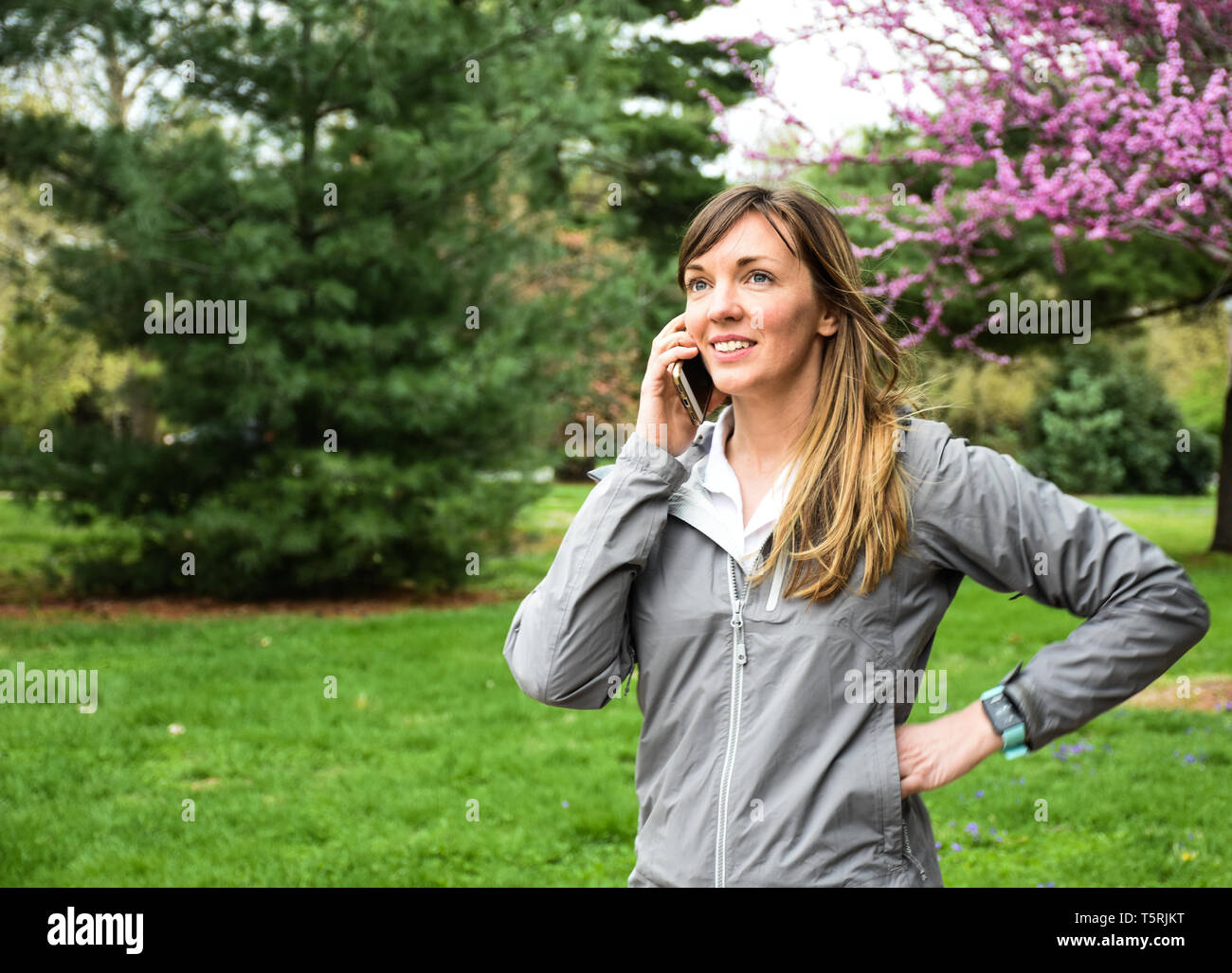 Attractive, smiling young woman talking on cell mobile phone, hands on hips, in city park, pink tree background, green grass, overcast day - Image Stock Photo