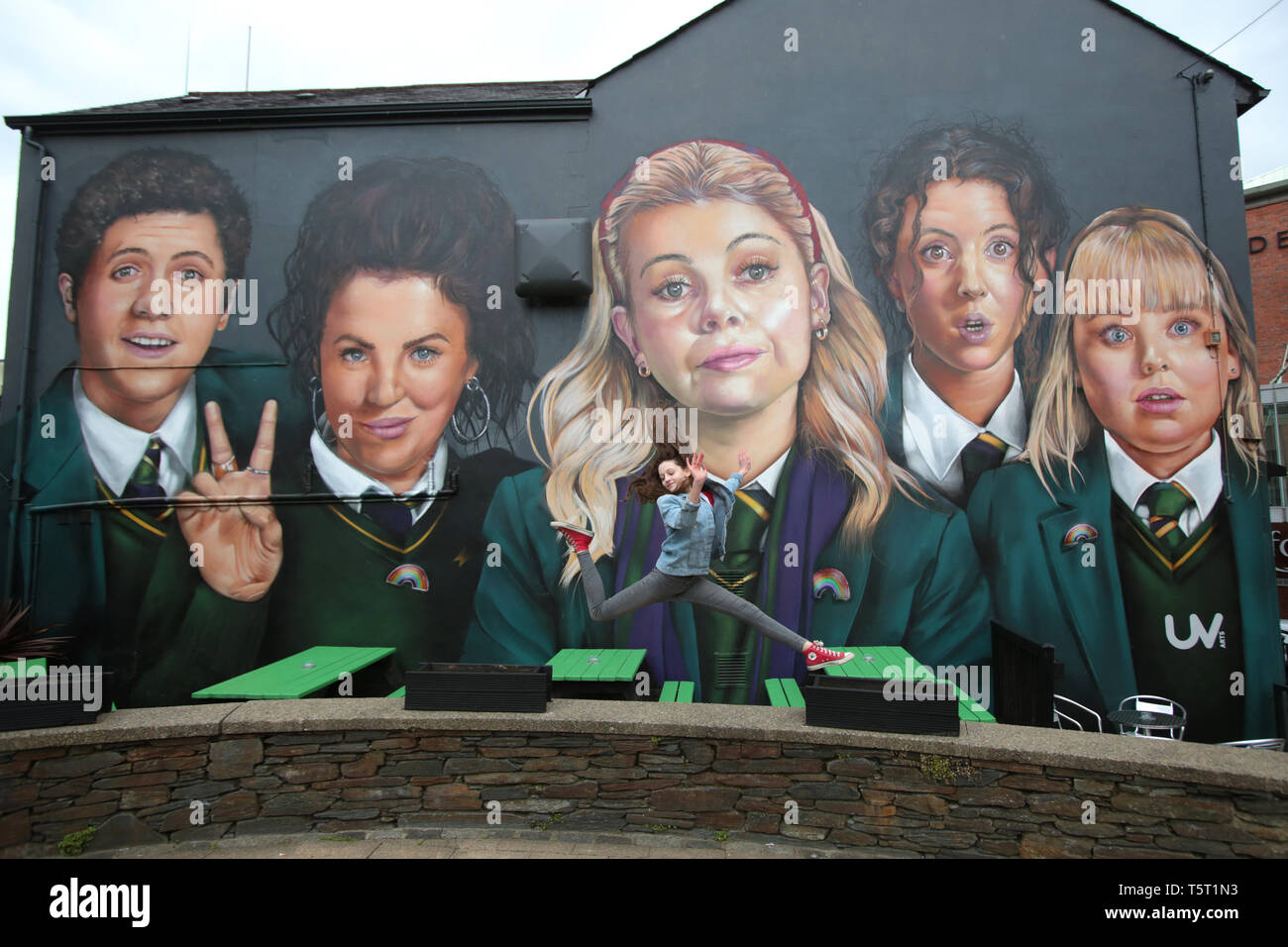 Derry, County Londonderry, Northern Ireland, 25th April, 2019 .A young girl jumps in front of a mural showing characters from the Channel 4 TV hit show Derry Girls in Derry/Londonderry, Northern Ireland. Derry Girls is a British television sitcom created and written by Lisa McGee. Staring Erin (Saoirse-Monica Jackson), her cousin Orla (Louisa Harland) and their friends Clare (Nicola Coughlan), Michelle (Jamie-Lee O'Donnell) and Michelle's English cousin James (Dylan Llewellyn) navigate their teen years during the Troubles in Derry, where they all attend a Catholic girls' secondary school. Erin Stock Photo
