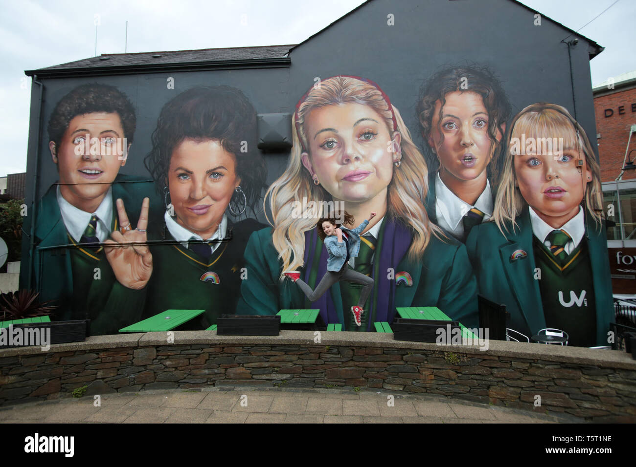 Derry, County Londonderry, Northern Ireland, 25th April, 2019 .A young girl jumps in front of a mural showing characters from the Channel 4 TV hit show Derry Girls in Derry/Londonderry, Northern Ireland. Derry Girls is a British television sitcom created and written by Lisa McGee. Staring Erin (Saoirse-Monica Jackson), her cousin Orla (Louisa Harland) and their friends Clare (Nicola Coughlan), Michelle (Jamie-Lee O'Donnell) and Michelle's English cousin James (Dylan Llewellyn) navigate their teen years during the Troubles in Derry, where they all attend a Catholic girls' secondary school. Erin Stock Photo