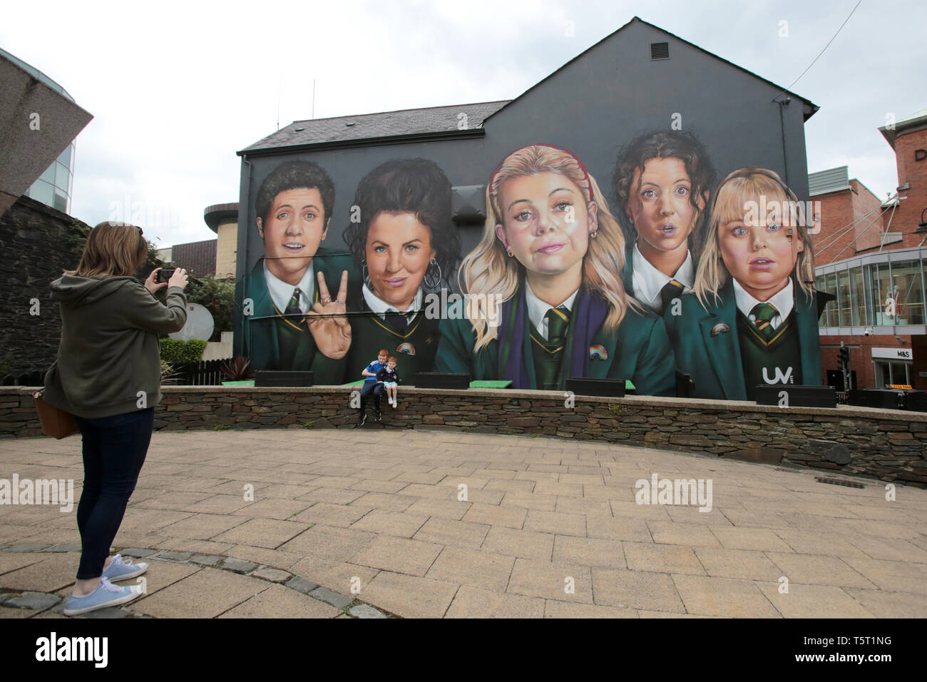Derry, County Londonderry, Northern Ireland, 25th April, 2019 .A family poses in front the Channel 4 hit TV show Derry Girls Mural in Derry/Londonderry, Northern Ireland. Derry Girls is a British television sitcom created and written by Lisa McGee. Staring Erin (Saoirse-Monica Jackson), her cousin Orla (Louisa Harland) and their friends Clare (Nicola Coughlan), Michelle (Jamie-Lee O'Donnell) and Michelle's English cousin James (Dylan Llewellyn) navigate their teen years during the Troubles in Derry, where they all attend a Catholic girls' secondary school. Erin lives with her father, Gerry; he Stock Photo