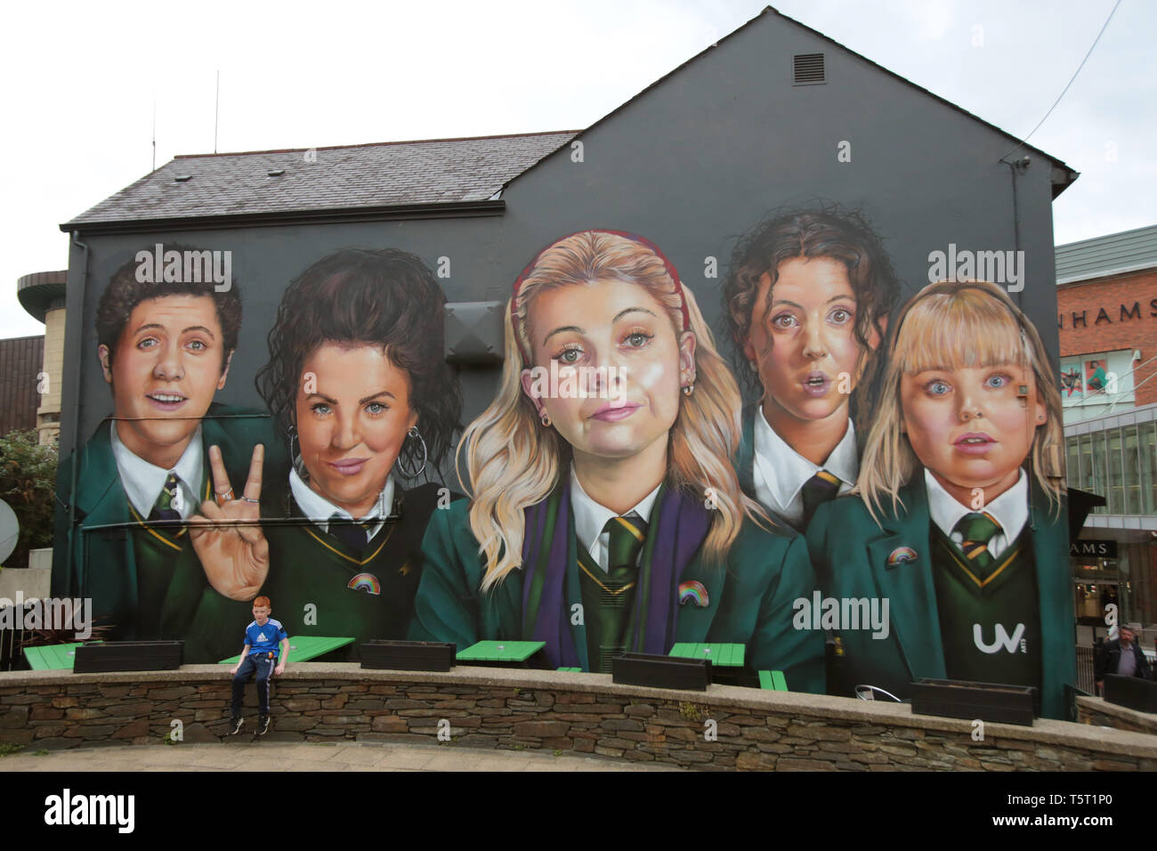 Derry, County Londonderry, Northern Ireland, 25th April, 2019 .A young boy sits on a wall in front of a mural showing characters from the Channel 4 TV hit show Derry Girls in Derry/Londonderry, Northern Ireland.  Derry Girls is a British television sitcom created and written by Lisa McGee. Staring Erin (Saoirse-Monica Jackson), her cousin Orla (Louisa Harland) and their friends Clare (Nicola Coughlan), Michelle (Jamie-Lee O'Donnell) and Michelle's English cousin James (Dylan Llewellyn) navigate their teen years during the Troubles in Derry, where they all attend a Catholic girls' secondary sch Stock Photo
