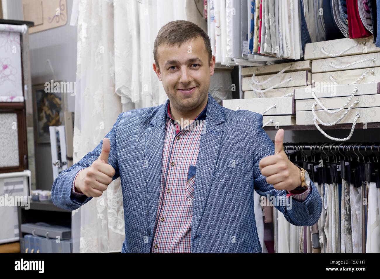 Young confident positive businessman fabric store owner showing thumbs up, gesture sign ok. Man standing in the store background interior fabric sampl Stock Photo