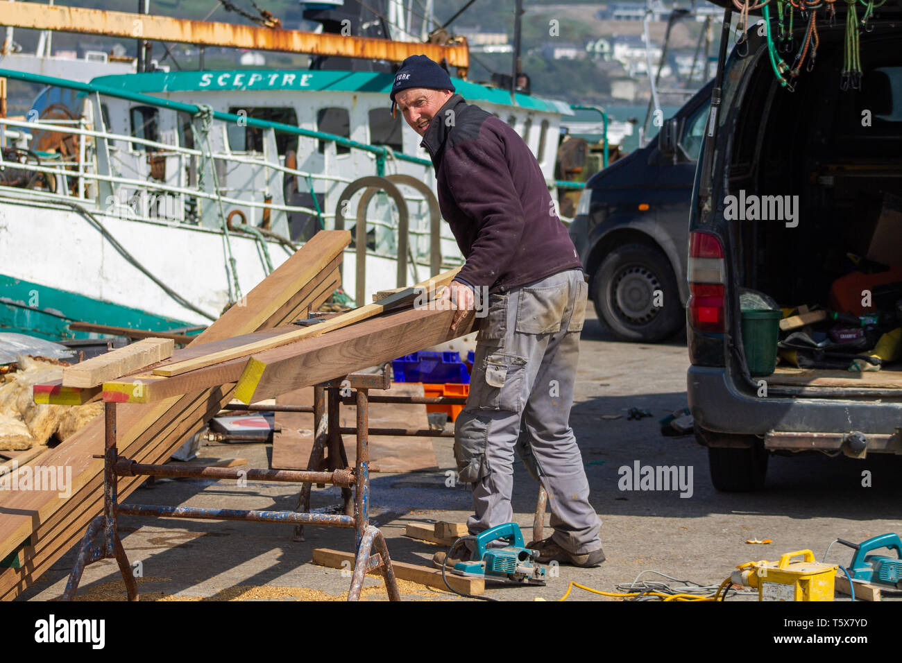 Carpenter shipwright selecting planks of teak wood from a pile of planks on the quayside. Stock Photo