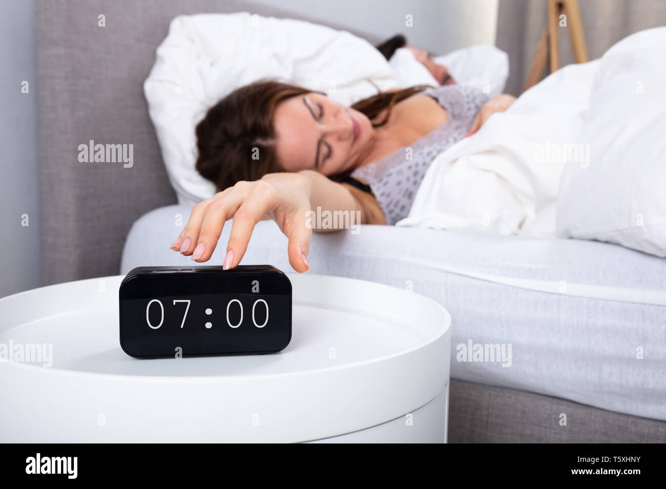 Young Woman Sleeping On Bed Turning Off Alarm Clock In Bedroom Stock