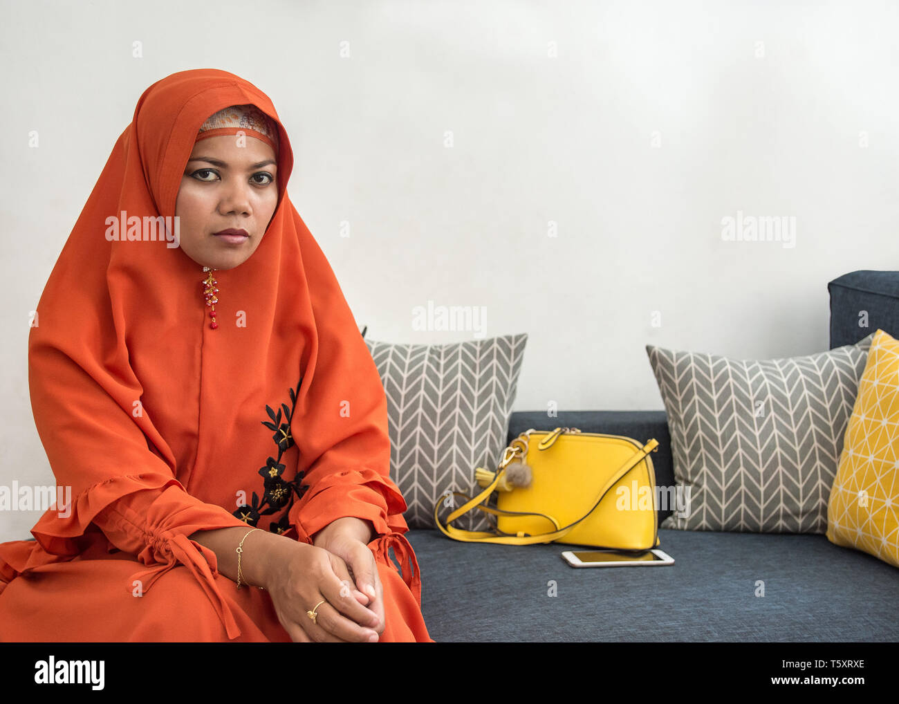 Asian woman wearing orange hijab outfit sitting on sofa looking at camera. Stock Photo