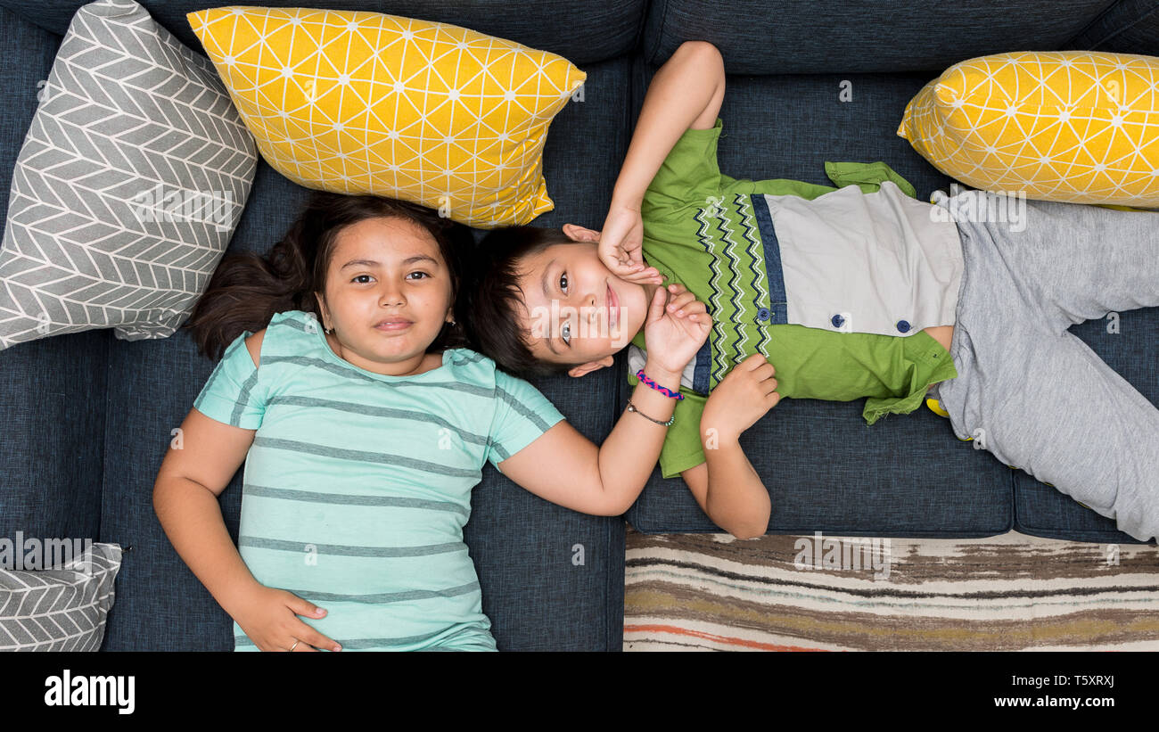 Mixed asian/caucasian brother and sister laying on sofa together smiling looking directly at camera. Stock Photo