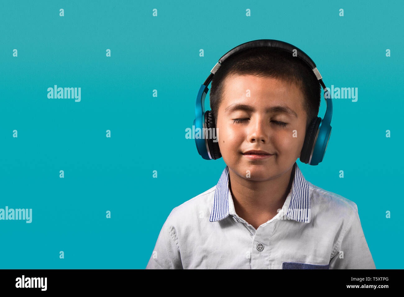 Young boy with closed eyes and smiling, happy wearing headphones listening to music close up portrait with blue background. Stock Photo