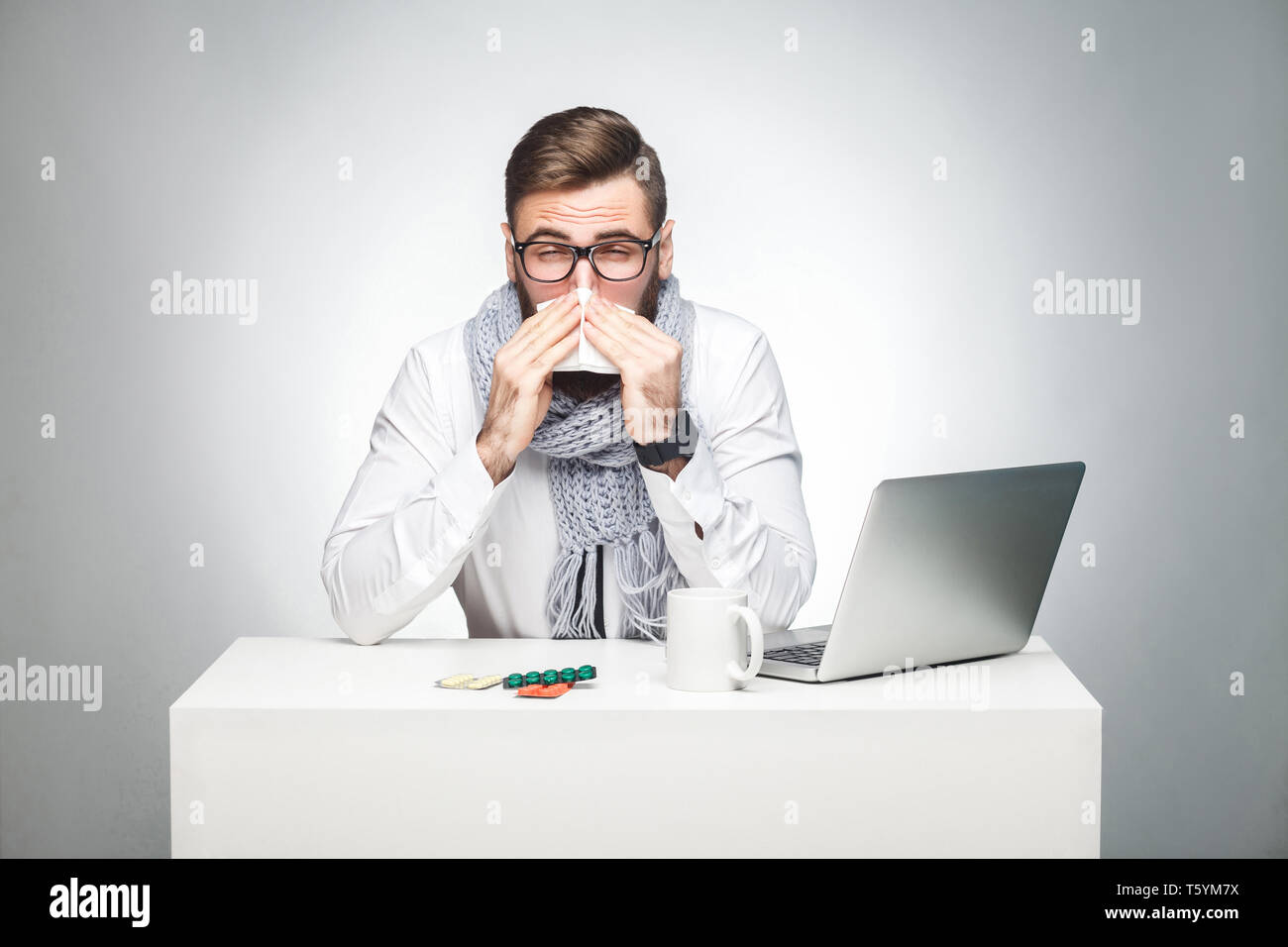Portrait of sick scribble young boss in white shirt, scarf and black tie are sitting in office and need to finish important report, have grippe virus. Stock Photo