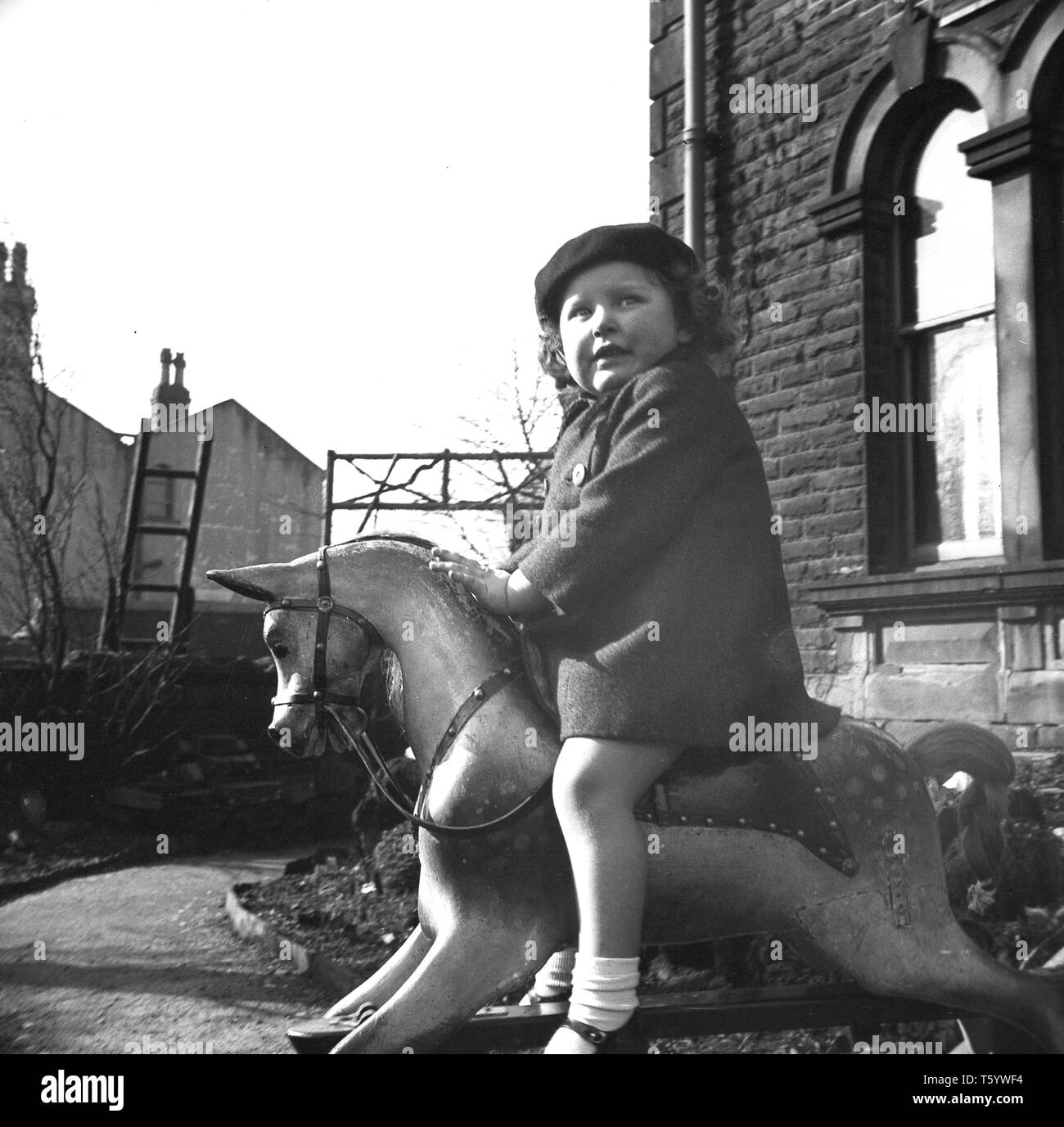1940s, historical, anxious little girl in a coat and berry riding on a rocking horse outside a house, England, UK. Stock Photo