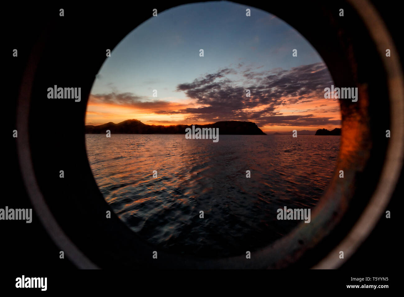 Komodo Island shoreline and spectacular colored sunset sky viewed through porthole window of fa boat Stock Photo