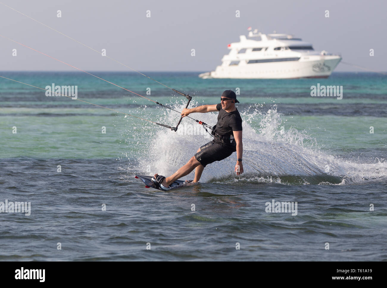 A kiteboarder wearing all black kicks up a huge spray in front of a large motor yacht. Stock Photo