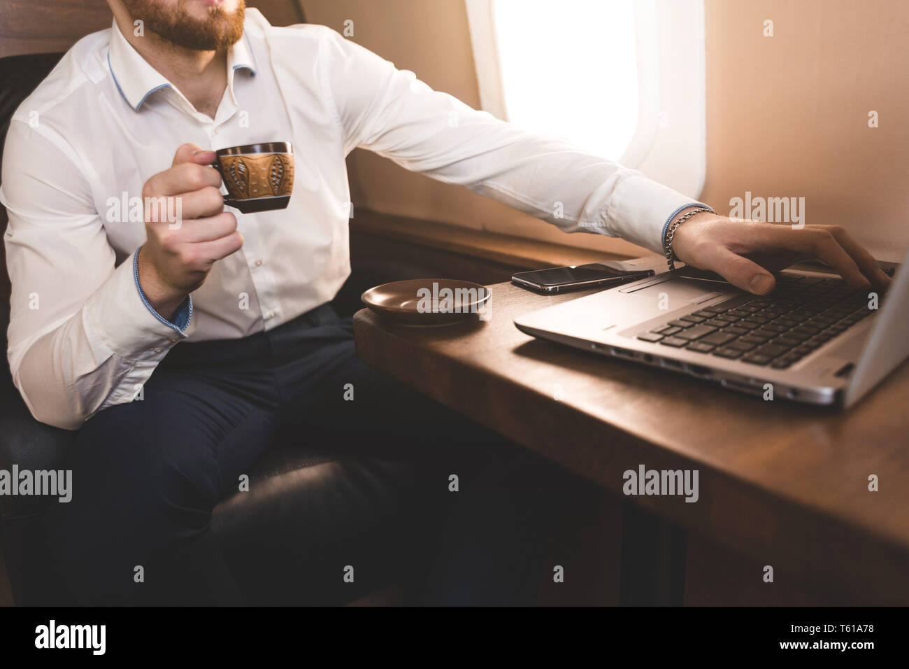 Attractive and successful businessman drinking coffee and working behind a laptop while sitting in a chair of his private jet. Stock Photo