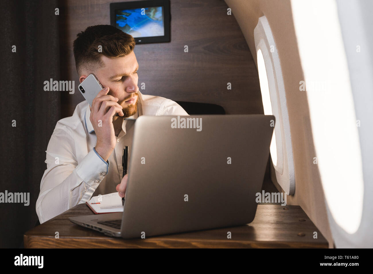 Young attractive and successful businessman talking on the phone and working while sitting in the chair of his private business plane Stock Photo