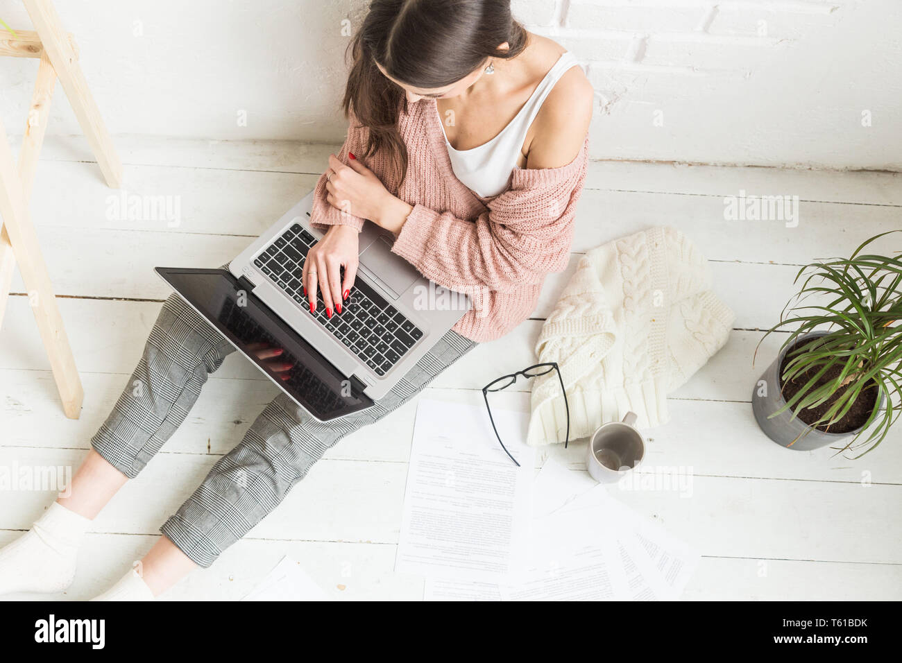 A young happy woman sits on the floor in a bright apartment or office interior and works at a laptop, freelancer girl at work, the concept of distance Stock Photo