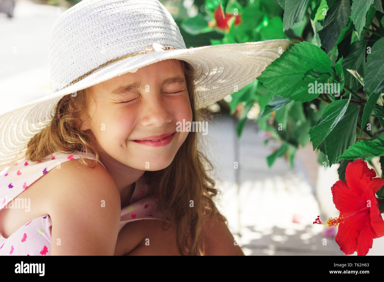 Beautiful little Girl looking at red flower and smiling in summer park. Happy cute kid playing outdoors. Stock Photo