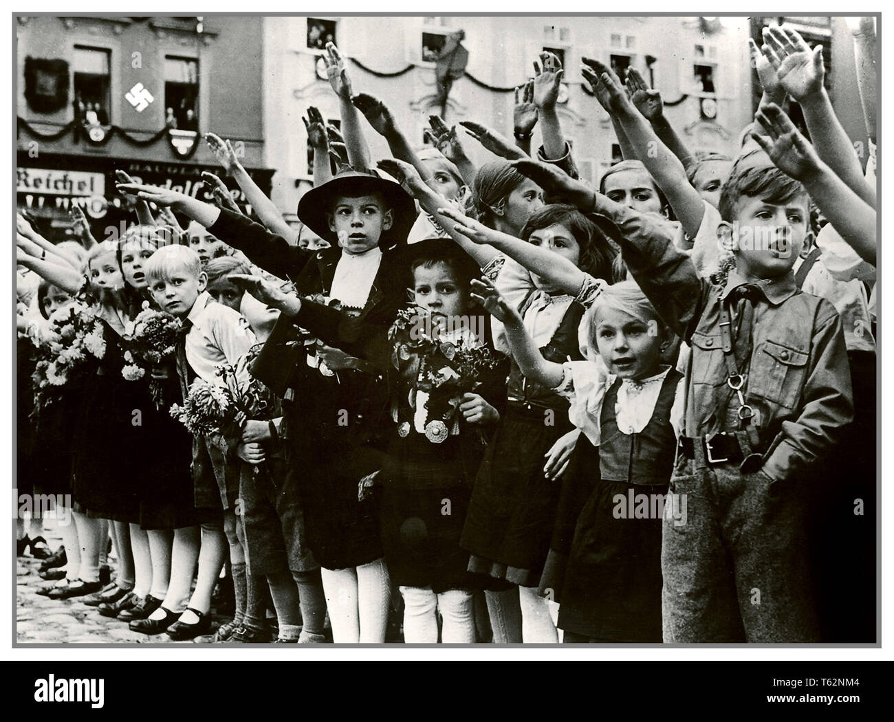 AUSTRIAN CHILDREN HEIL HITLER SALUTE 1930's Nazi Propaganda image of Austrian children group welcoming Adolf Hitler with traditional Heil Hitler salute with arms raised. Swastika symbol on wall in background Sudetenland Stock Photo