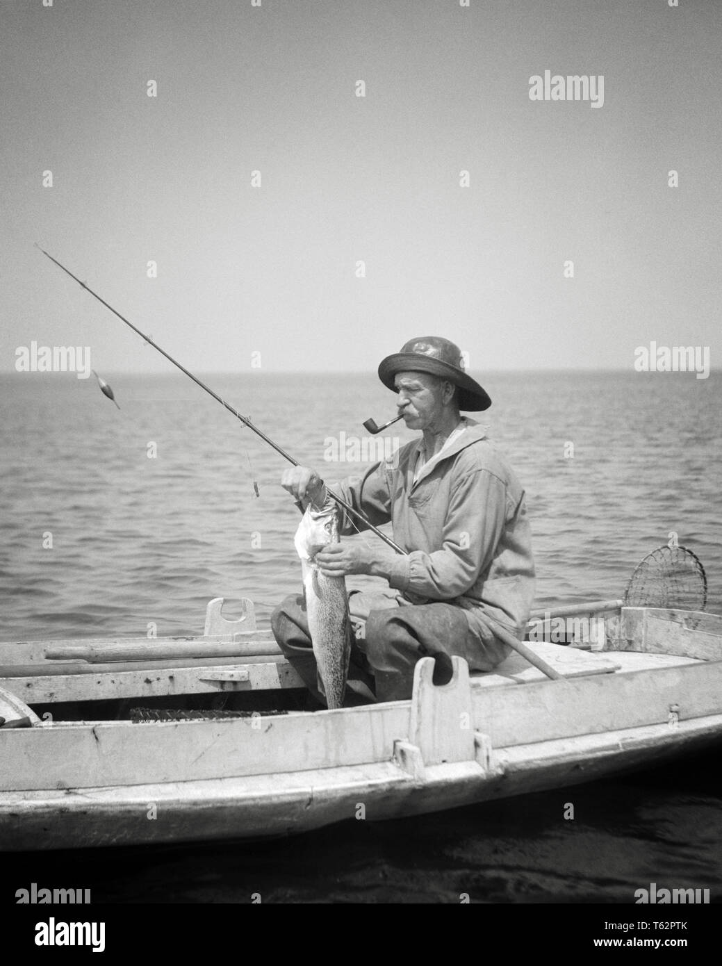 1920s MAN BARNEGAT BAY COMMERCIAL FISHERMAN SITTING IN DUCK BOAT SMOKING PIPE REMOVING FISHHOOK FROM FISH CATCH NEW JERSEY USA - a143 HAR001 HARS B&W MIDDLE-AGED MAN SUCCESS SKILL ACTIVITY AMUSEMENT CANVAS HOBBY INTEREST REMOVING HOBBIES KNOWLEDGE PASTIME PLEASURE NJ OCCUPATIONS DUCK BOAT FISHING ROD NEW JERSEY OLD SALT WAXED OILSKIN RELAXATION AMATEUR BARNEGAT BAY BLACK AND WHITE CAUCASIAN ETHNICITY ENJOYMENT HAR001 OLD FASHIONED Stock Photo
