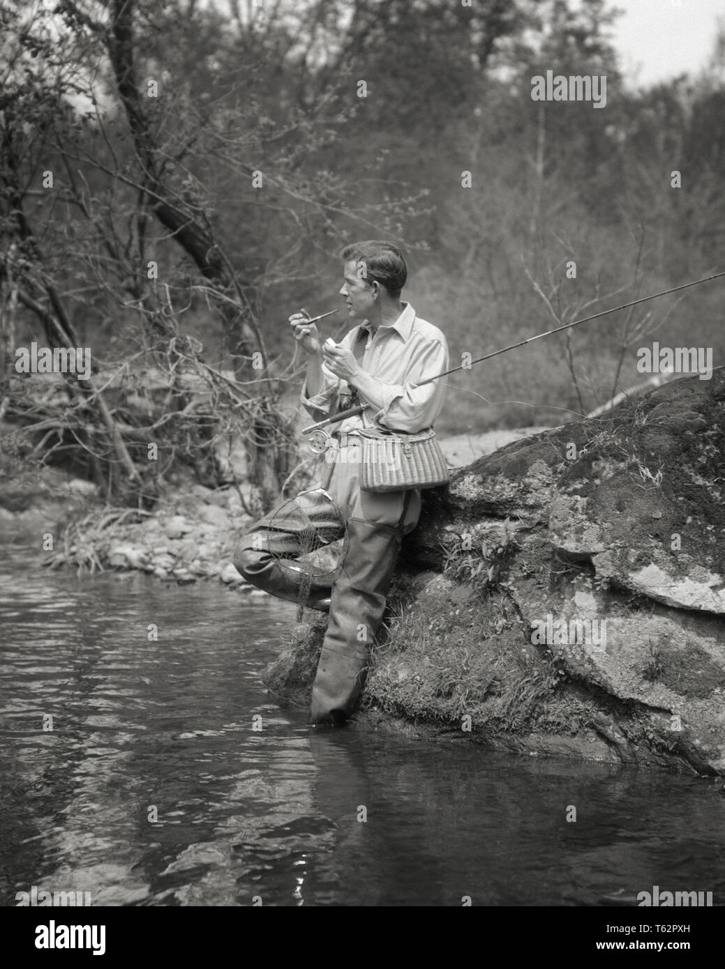 1920s FLY FISHERMAN WEARING WADERS LIGHTING PIPE TAKING A BREAK LEANING ON BOULDER ON BANK OF STREAM - a3361 HAR001 HARS TOBACCO INTEREST HOBBIES KNOWLEDGE RECREATION PASTIME REEL PLEASURE ESCAPE STYLISH WADERS ANGLING MID-ADULT MID-ADULT MAN RELAXATION AMATEUR BLACK AND WHITE BOULDER CAUCASIAN ETHNICITY CREEL ENJOYMENT HAR001 OLD FASHIONED Stock Photo