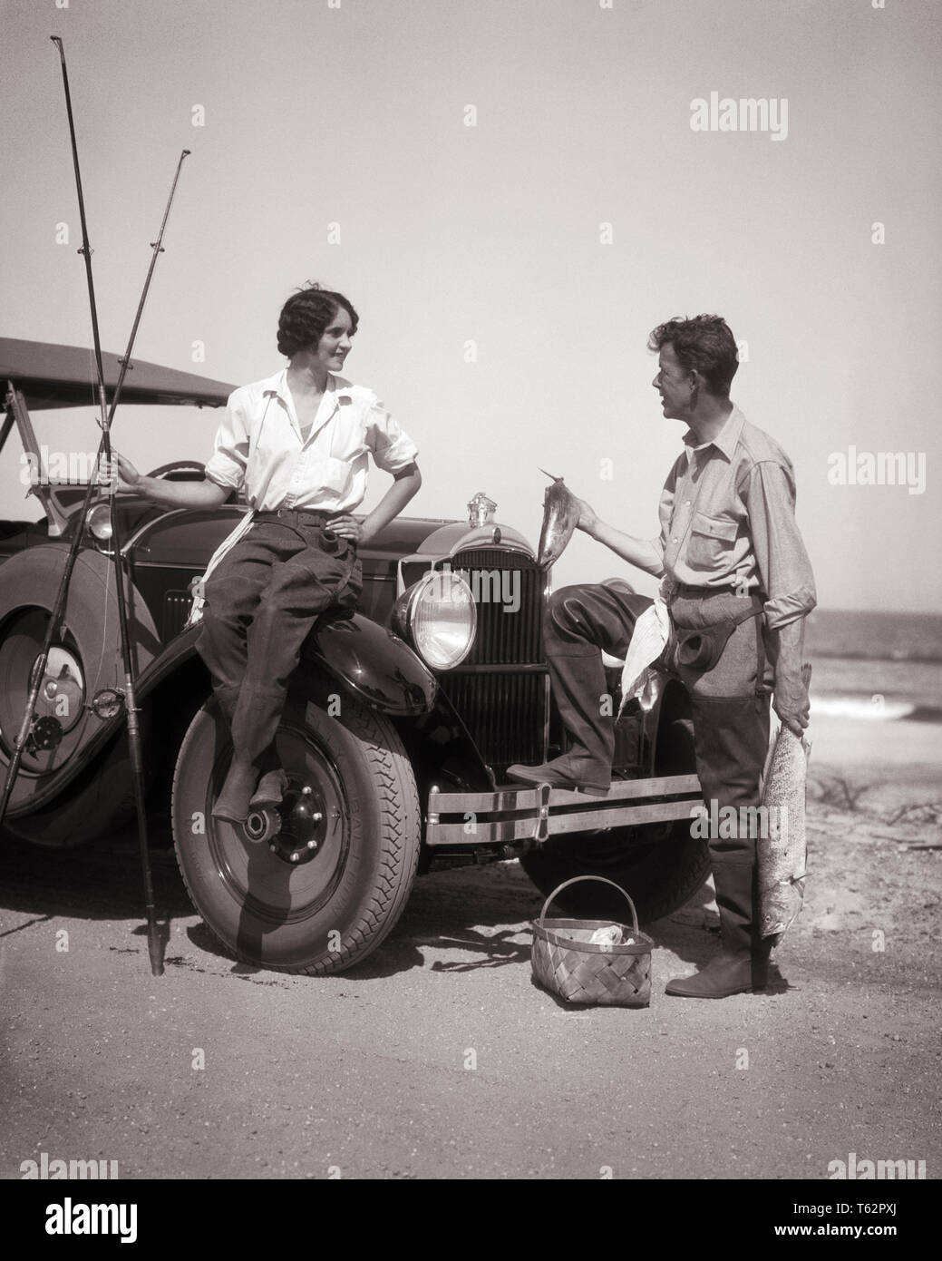 1920s COUPLE SURF FISHING MAN STANDING HOLDING CATCH WOMAN SITTING ON CAR FENDER HOLDING RODS - a3555 HAR001 HARS TRANSPORTATION B&W GOALS HAPPINESS ADVENTURE AUTOS RECREATION AUTOMOBILES VEHICLES ANGLING FENDER MID-ADULT MID-ADULT MAN RODS TOGETHERNESS YOUNG ADULT WOMAN BLACK AND WHITE CAUCASIAN ETHNICITY HAR001 OLD FASHIONED Stock Photo