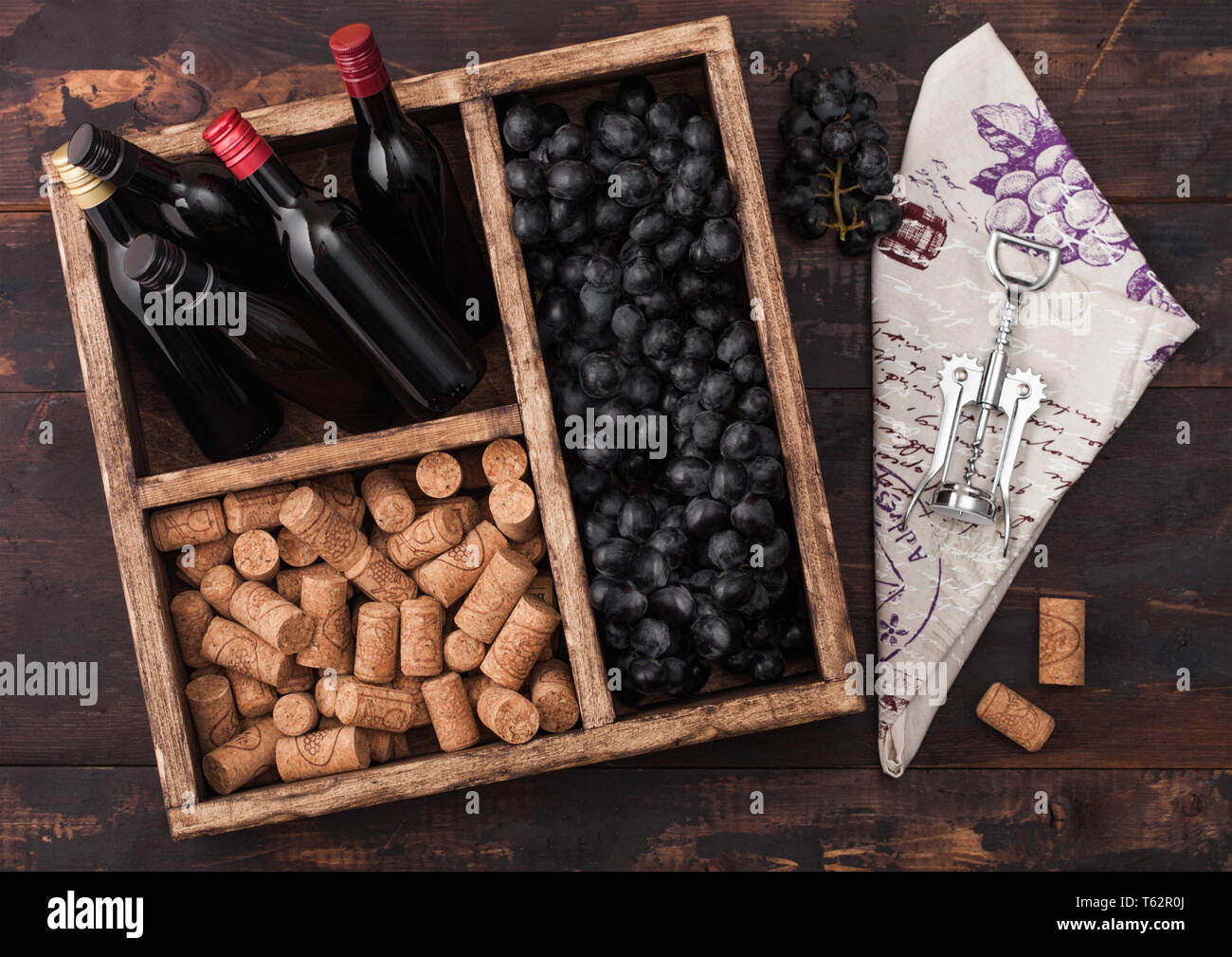 Mini bottles of red wine with dark grapes with corks and corkscrew inside vintage wooden box on dark wooden background with linen towel. Stock Photo