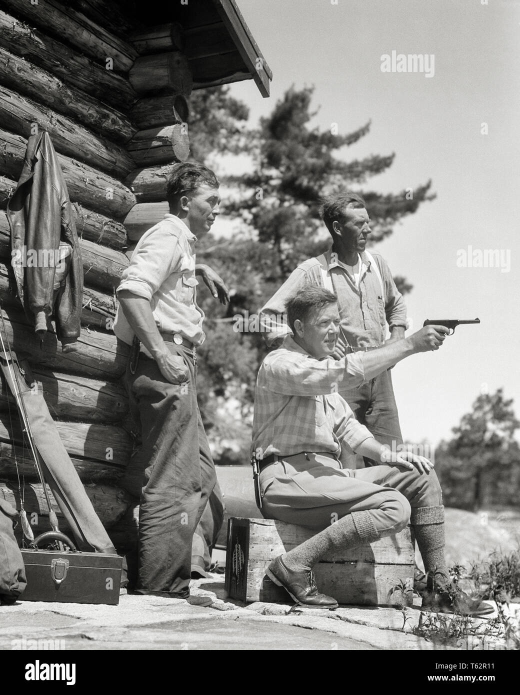 1930s THREE MEN TOGETHER OUTSIDE NORTH WOODS HUNTING FISHING LOG CABIN SEATED MAN AIMING PLINKING WITH COLT 22 CALIBER PISTOL - a5529 HAR001 HARS ADVENTURE LEISURE LOG EXTERIOR RECREATION CONCEPTUAL STYLISH SPORTSMEN AIMING COLT FIREARM FIREARMS HUNTERS MID-ADULT MID-ADULT MAN PISTOL RELAXATION TOGETHERNESS 22 CALIBER BLACK AND WHITE CAUCASIAN ETHNICITY HAR001 OLD FASHIONED Stock Photo