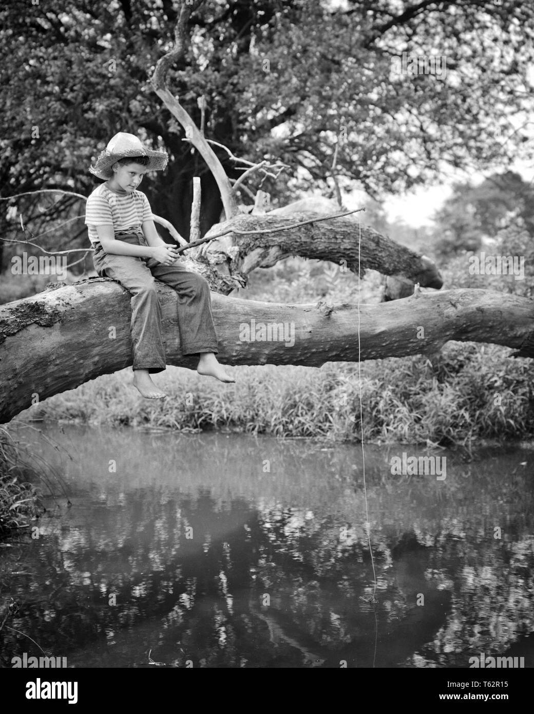1940s BAREFOOT COUNTRY BOY WEARING BLUE JEANS STRIPED SHIRT AND STRAW HAT SITTING ON A FALLEN TREE OVER A CREEK FISHING  - a64 HAR001 HARS SERENITY B&W SKILL ACTIVITY AMUSEMENT ADVENTURE FALLEN HOBBY LEISURE INTEREST AND HOBBIES KNOWLEDGE RECREATION PASTIME PLEASURE ON BAREFOOT STRAW HAT HUCK FINN T-SHIRT TOM SAWYER BLUE JEANS HUCKLEBERRY FINN JUVENILES RELAXATION AMATEUR BLACK AND WHITE CAUCASIAN ETHNICITY CREEK ENJOYMENT HAR001 OLD FASHIONED Stock Photo