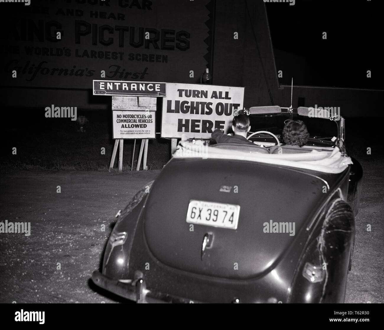 1940s COUPLE MAN WOMAN IN CONVERTIBLE CAR PULLING IN TO DRIVE-IN MOVIE THEATER ENTRANCE SIGN - asp ap 8925 ASP001 HARS TEENAGE BOY ENTERTAINMENT B&W MOVIES DATING DREAMS ENTERING IN TO ATTRACTION COURTSHIP ESCAPE DATE NIGHT MOTION PICTURES POSSIBILITY DRIVE IN DRIVE-IN NIGHTTIME REARVIEW SOCIAL ACTIVITY TOGETHERNESS YOUNG ADULT MAN YOUNG ADULT WOMAN 1948 BLACK AND WHITE CAUCASIAN ETHNICITY COURTING OLD FASHIONED Stock Photo