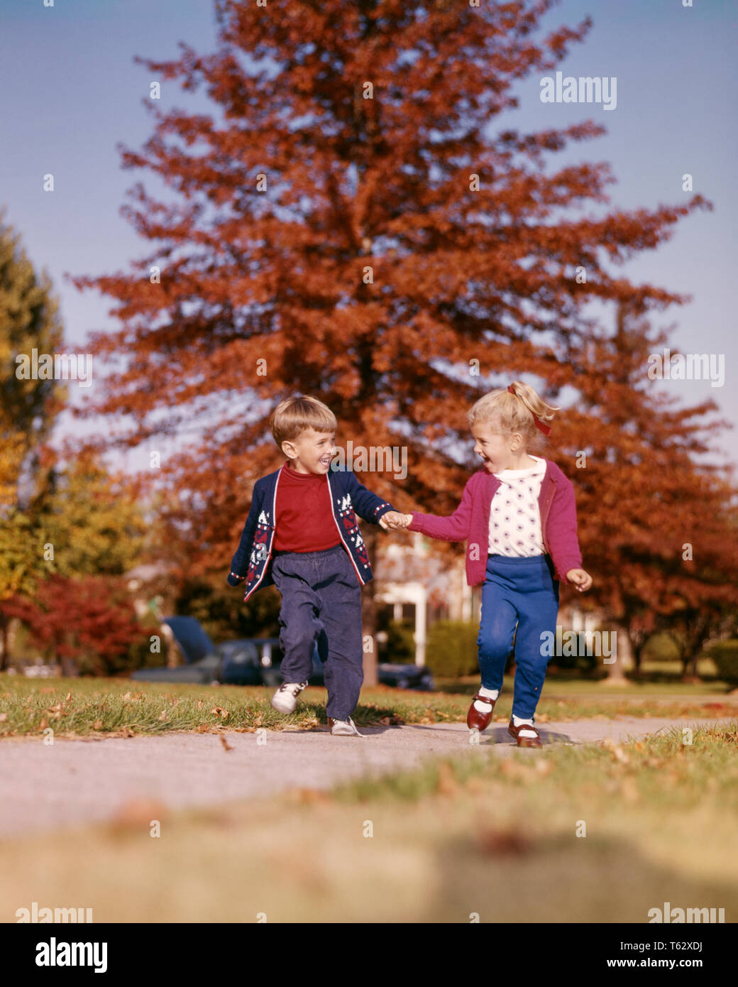 1960s SMILING BOY AND GIRL RUNNING SKIPPING HOLDING HANDS ON SIDEWALK AUTUMN  - kj3562 HAR001 HARS BROTHERS HEALTHINESS HOME LIFE NATURE COPY SPACE FRIENDSHIP FULL-LENGTH MALES SIBLINGS SISTERS HAPPINESS ADVENTURE AND LOW ANGLE RECREATION SKIPPING FALL SEASON HOLDING HANDS SIBLING CONNECTION ESCAPE FRIENDLY STYLISH JUVENILES MARY JANE SHOES TOGETHERNESS AUTUMNAL CAUCASIAN ETHNICITY FALL FOLIAGE HAR001 OLD FASHIONED Stock Photo