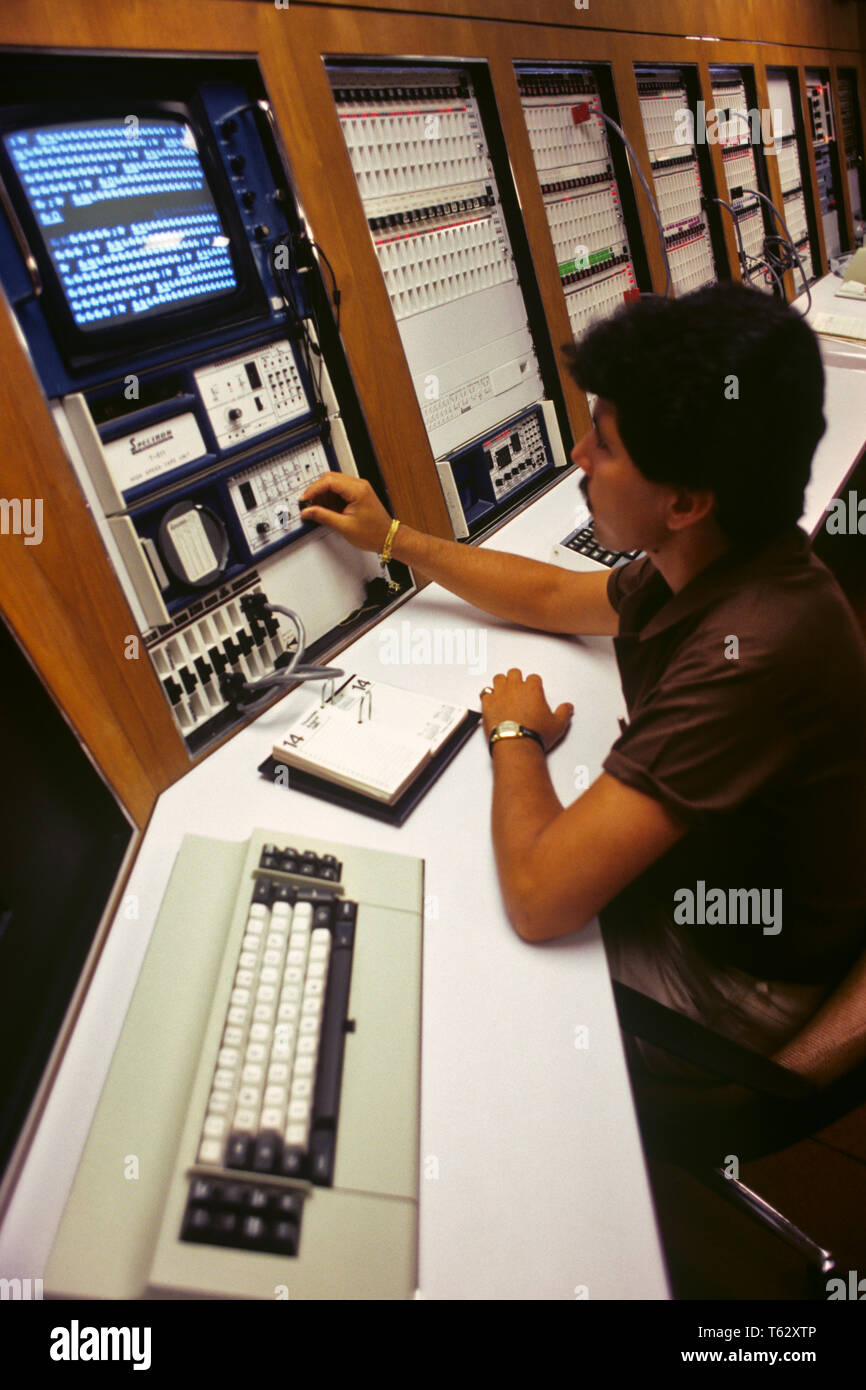 1980s AFRICAN AMERICAN MAN TECHNICIAN  WORKING AT COMPUTER MAINFRAME MONITOR   - ko1316 PHT001 HARS HIGH ANGLE SCIENTIFIC AFRICAN-AMERICANS AFRICAN-AMERICAN PROGRESS BLACK ETHNICITY INNOVATION EMPLOYMENT OCCUPATIONS HIGH TECH CONNECTION KEYPAD COMPLEX COMPLICATED MONITOR SWITCHES EMPLOYEE YOUNG ADULT MAN HISPANIC ETHNICITY OLD FASHIONED AFRICAN AMERICANS Stock Photo