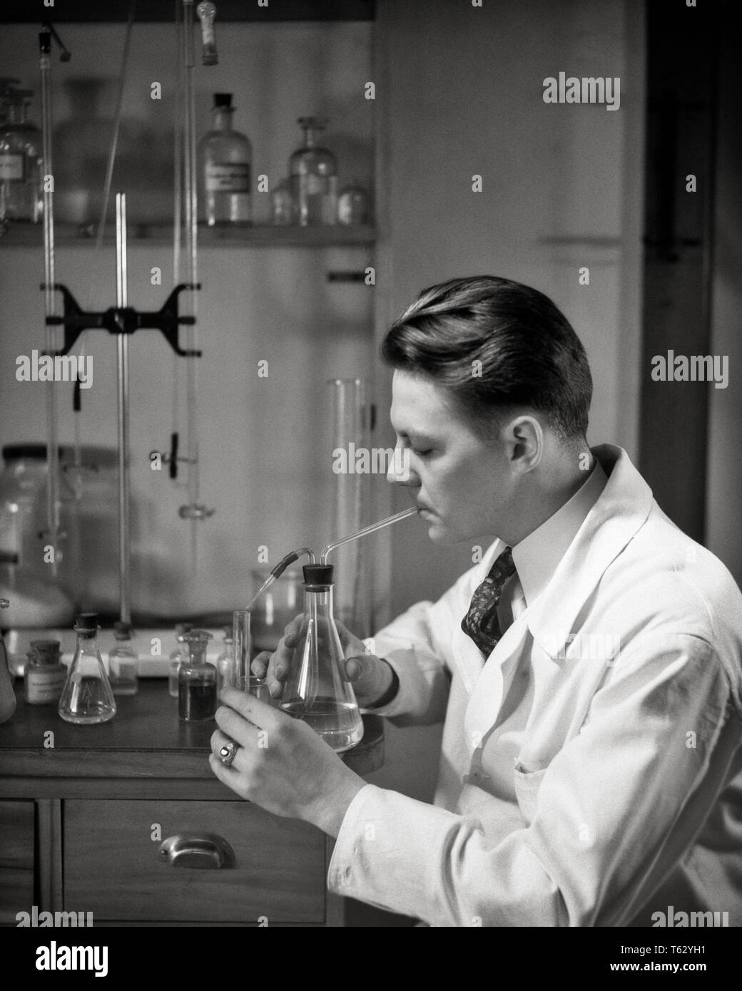 1930s MAN WORKING IN SCIENCE LABORATORY PIPETTING LIQUID FROM FLASK INTO A TEST TUBE - l1842 HAR001 HARS SKILL OCCUPATION SKILLS DISCOVERY CHEMIST SCIENTIFIC INNOVATION LABOR EMPLOYMENT OCCUPATIONS CONNECTION EMPLOYEE FLASK SOLUTIONS YOUNG ADULT MAN BLACK AND WHITE CAUCASIAN ETHNICITY CHEMICALS HAR001 LABORATORIES LABORING LABS OLD FASHIONED Stock Photo